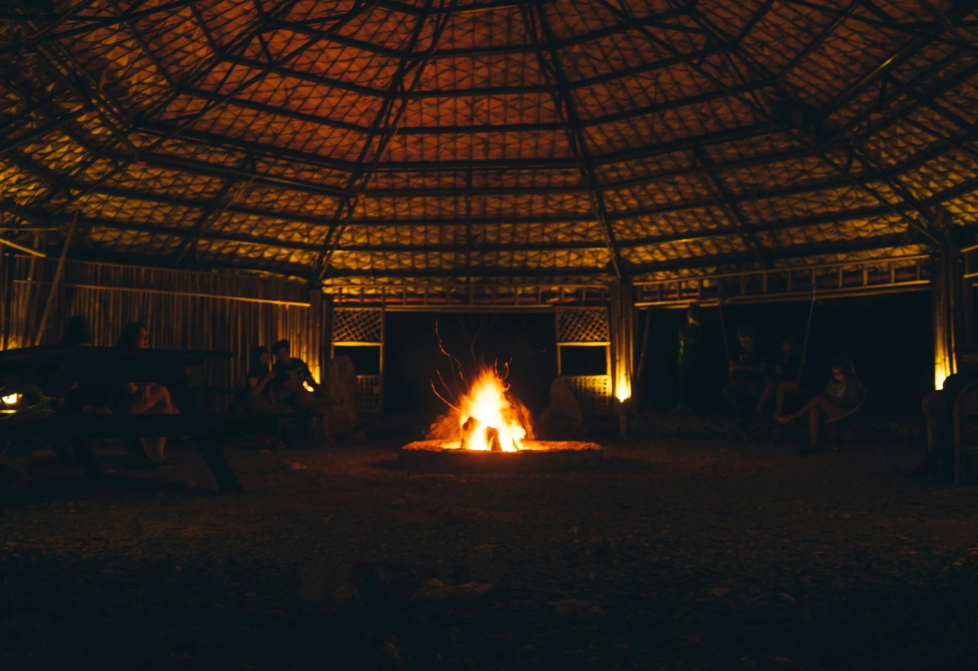A fire burning in the middle of a round thatched building at night, people sitting around the edge in the dark.